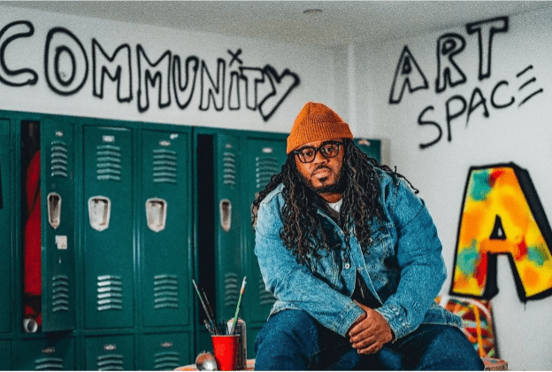 A photo of Cue Perry in a room with spray paint art behind him. 